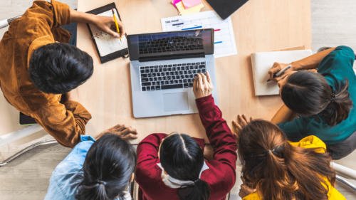 Um gripo de cinco mulheres está em uma mesa diante de um laptop, uma delas toca no teclado do laptop enquanto outra toma notas